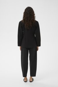 Svart skräddarsydd blazer med markerade axlar, sedd bakifrån, kombinerad med lösa svarta byxor och svarta platta sandaler. Mjukt tyg.