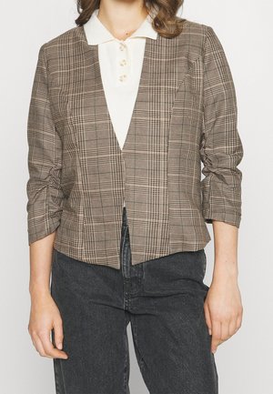 Kvinde iført en brun ternet blazer med trekvartlange ærmer over en hvid skjorte med krave og mørkegrå jeans.