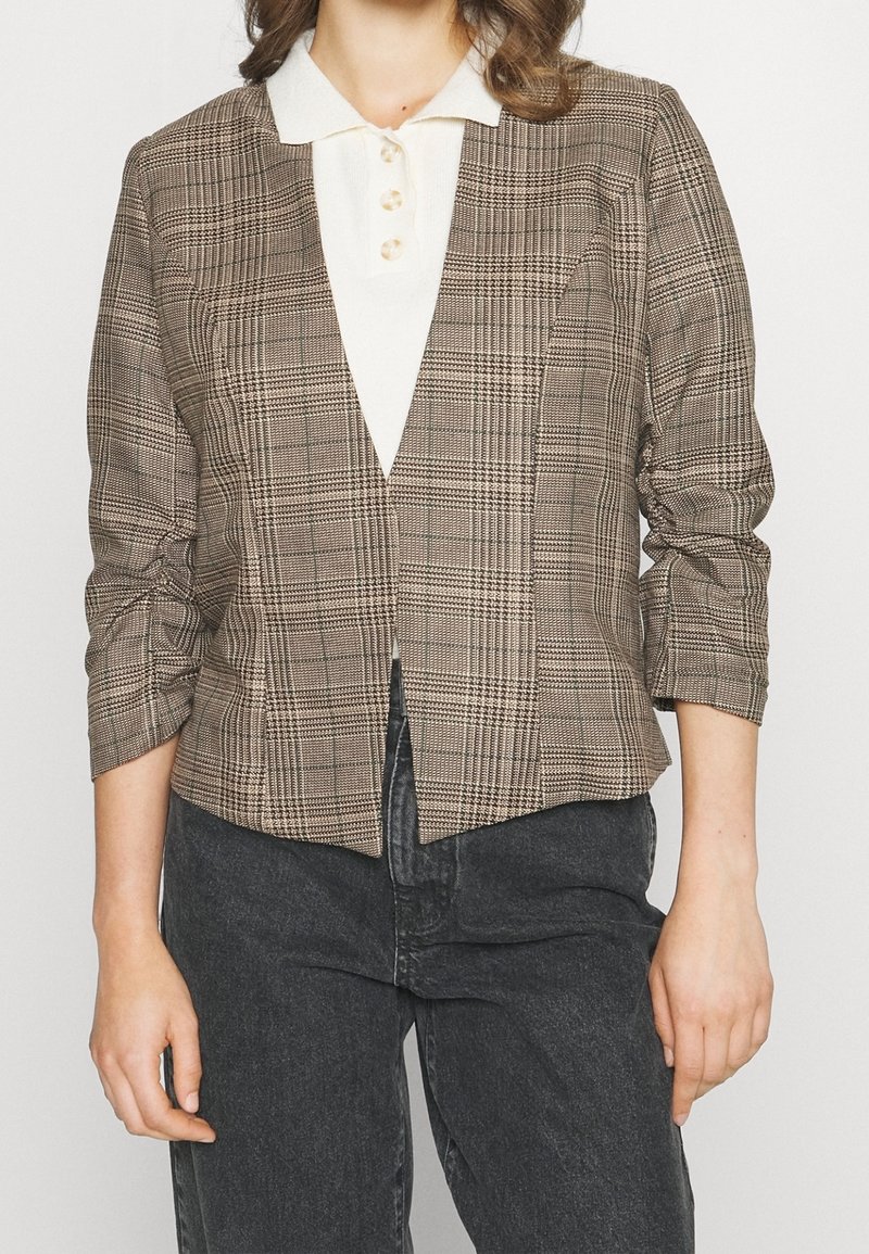 Femme portant un blazer marron à carreaux à manches trois-quarts sur une chemise blanche boutonnée avec col et un jean gris foncé.