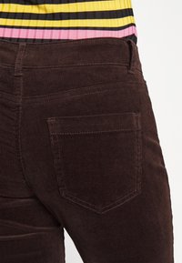 Gros plan sur une personne portant un pantalon en velours côtelé marron foncé avec une poche arrière et un haut rayé jaune, rose et noir.