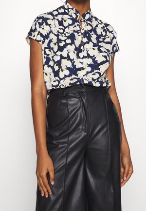 Kobieta ubrana w krótką bluzkę w kwiaty z wycięciem przy dekolcie i czarne, wysokie skórzane spodnie, stojąca na jednolitym tle.