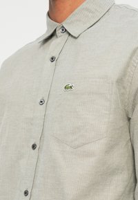Camicia button-up grigio chiaro con texture a spina di pesce, dotata di un piccolo logo ricamato di un coccodrillo verde e taschino sul petto con alette appuntite.