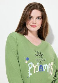 Top verde de manga larga con escote en V, que presenta el texto "La vida es mejor en PIJAMAS" en varios colores y gráficos de flores. Textura de tela suave.