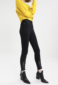 Černé džíny s vysokým pasem a dekorativními šněrovacími detaily po stranách, v kombinaci s žlutým teksturovaným svetrem a černými kozačkami do kotníků.