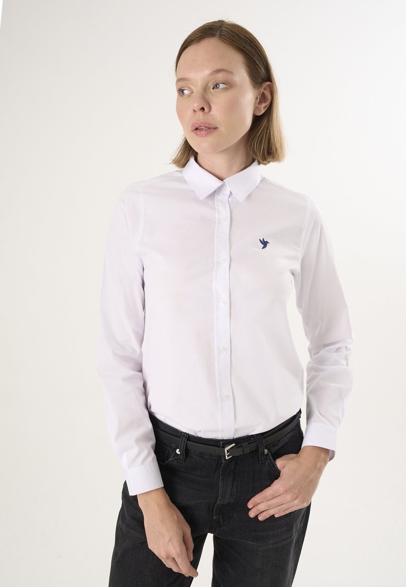 Camicia bianca a maniche lunghe con collo, con un piccolo logo di un uccellino blu sul lato sinistro del petto. Indossata con jeans neri e una cintura nera.