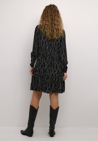 Abito nero a fantasia con maniche lunghe, orlo arricciato e vestibilità ampia. Abbinato a stivali neri fino al ginocchio. Texture liscia e design fluido.