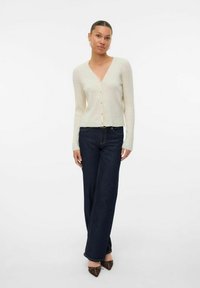 Ljus beige ribbad cardigan med V-halsringning, långa ärmar och knäppning, i kombination med mörkblå vida jeans och bruna spetsiga skor.