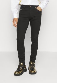 Černé džíny skinny z denimu s hladkou texturou a zlatými kovovými doplňky; kombinované s lesklými černými a zlatými teniskami.