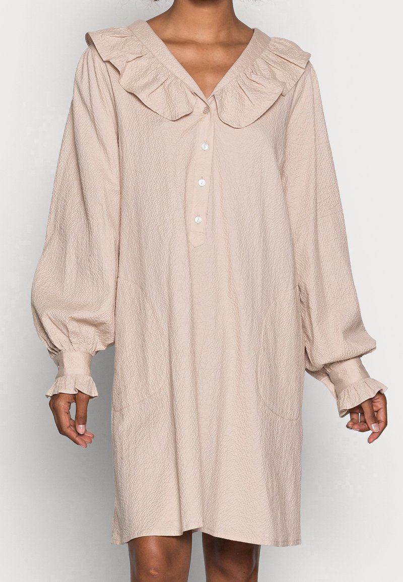 Vestido beige pálido con un tejido texturizado, gran cuello con volantes, cierre frontal de botones y mangas largas abullonadas con puños con volantes.