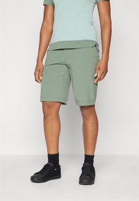 Pantalones cortos verdes de tela suave, hasta la rodilla con dobladillos enrollados. Combinados con una camiseta de color azul claro y zapatillas negras.