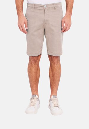 Uomo che indossa pantaloncini beige fino al ginocchio, scarpe da ginnastica bianche e una maglietta bianca, in piedi davanti a uno sfondo chiaro e uniforme.