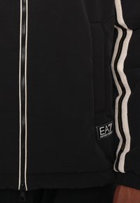 Črna puhovka z zadrgo spredaj, belimi in zlatimi črtami na rokavih, teksturiranim materialom ter označeno znamko na spodnjem levem robu.