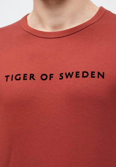 Crvena pamučna majica s okruglim izrezom i hrabrim crnim tekstom "TIGER OF SWEDEN" smještenim centralno na prednjoj strani.