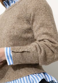 Pull en laine marron avec une texture côtelée et des détails à trois boutons sur les poignets, superposé sur une chemise à rayures bleues et blanches.