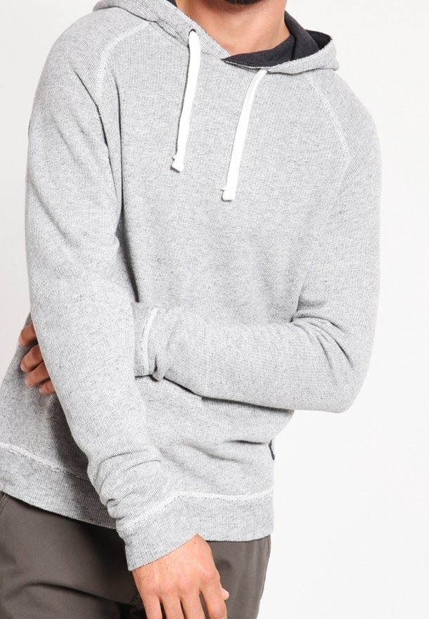 Homme portant un sweat à capuche gris clair avec des cordons blancs et un pantalon foncé, bras croisés devant un fond blanc.