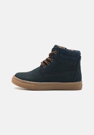 Bottines en daim bleu marine avec semelle en caoutchouc brun, bout rond et lacets marron. Comprend un col rembourré et des coutures décoratives.