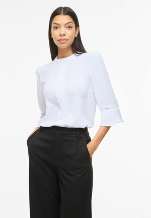 Kobieta z długimi ciemnymi włosami, ubrana w białą bluzkę z rękawami 3/4 i czarne spodnie z wysokim stanem, stoi z rękami w kieszeniach.