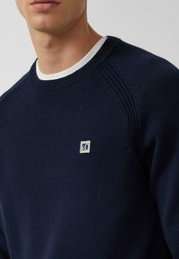 Mężczyzna w granatowym swetrze z dzianiny nosi biały t-shirt z okrągłym dekoltem, z małym białym prostokątnym logo na lewej piersi.