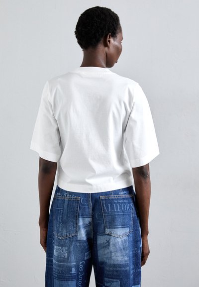 White short-sleeved cotton t-shirt with a crew neck, paired with blue denim jeans featuring printed patterns and patch pockets.