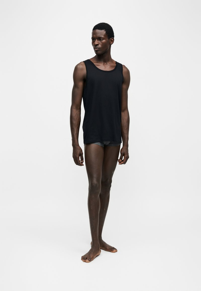 Homme grand à la peau foncée portant un débardeur noir et un short foncé, debout pieds nus sur un fond clair uni.