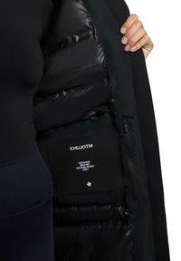 Jachetă tip puffer cu interior lucios, de culoare neagră și exterior mat; dispune de un buzunar cu fermoar având o etichetă pe care scrie "KHUJOTM" și detalii ale mărcii.