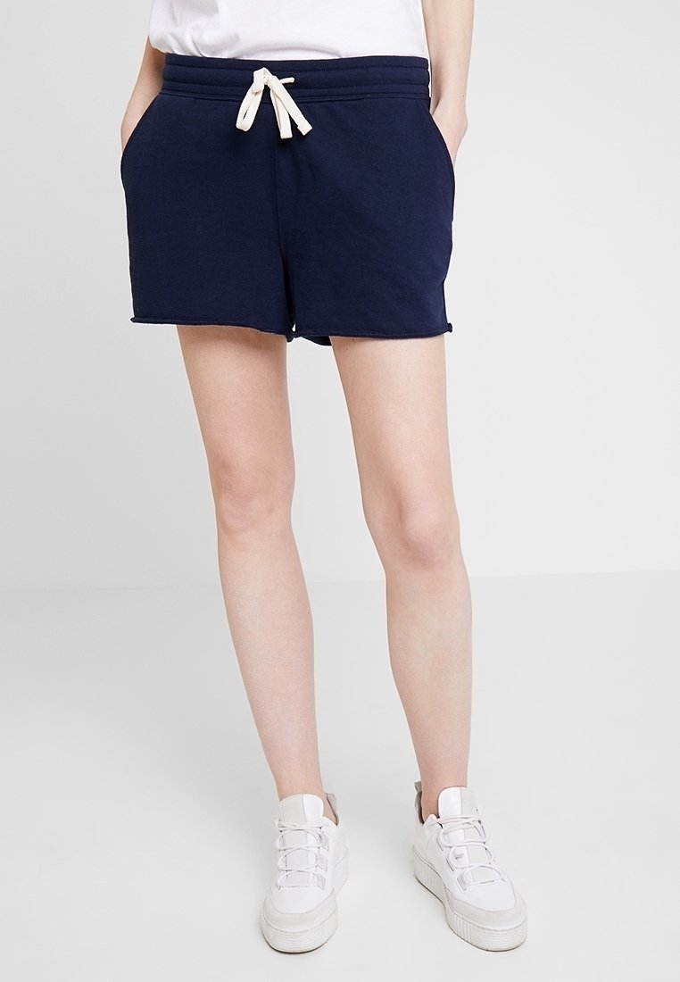 Personne portant un short bleu marine avec cordon et des baskets blanches, se tenant devant un fond gris clair uni.