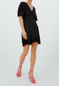 Nainen, jolla on päällään lyhyt musta kietaisumekko puffihihnoilla ja vaaleanpunaiset remmisandaalit korkokengillä, seisoo yksinkertaista vaaleaa taustaa vasten.