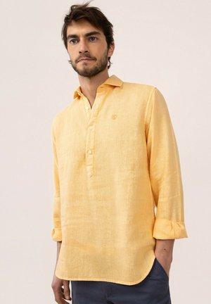 Hombre con el pelo corto y oscuro y barba, que lleva una camisa de lino de manga larga de color amarillo claro con las mangas remangadas y pantalones oscuros.