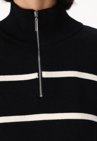 Pull en maille noire avec col montant et demi-fermeture éclair, orné de rayures horizontales blanches. Tissu texturé avec une finition douce.