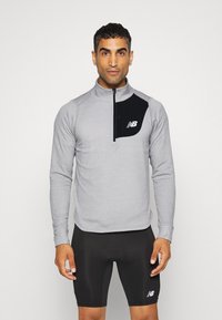 Camisola atlética de manga longa, cinza claro, com gola de meia-zíper, padrão texturizado e um painel preto no peito com um logótipo branco. Combinada com calções pretos.
