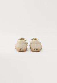 Beige suedesko med lavt design, gummisåler og en logodetalj på hælen. Parret står plassert rygg mot rygg.