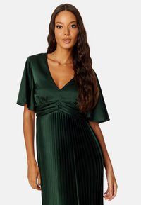 Robe en satin vert foncé avec décolleté en V, manches courtes volantées et jupe plissée. Présente un ruché à la taille pour plus de texture.