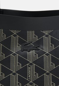 Matériau noir texturé avec des lignes géométriques dorées, un logo crocodile noir embossé et le mot Lacoste en lettres dorées.
