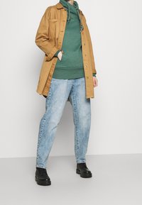 Cappotto lungo tan sopra una felpa verde con cappuccio, abbinato a jeans azzurri larghi e stivaletti neri alla caviglia. Outfit casual senza motivi visibili.