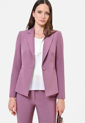 Femme portant un costume mauve sur mesure avec un pantalon assorti et un chemisier blanc à volants, tenant un petit sac à main sombre dans sa main gauche.