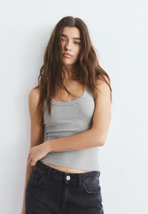 Fiatal nő hosszú barna hajjal, szürke ujjatlan felsőt és fekete farmert visel, egyszerű fehér háttér előtt áll.