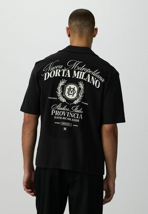 Mann, der mit dem Rücken zur Kamera steht, trägt ein schwarzes Kurzarmhemd mit weißem italienischem Text und Wappen-Design auf der Rückseite.