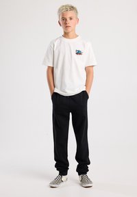 White short-sleeve T-shirt with colorful graphic on left chest, paired with black sweatpants and checkerboard slip-on shoes.