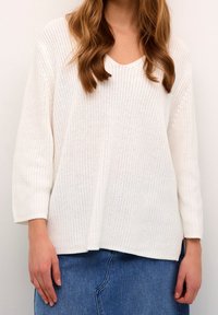 Femme portant un pull en maille blanc à manches trois-quarts et une jupe en denim bleu, debout devant un fond clair uni.