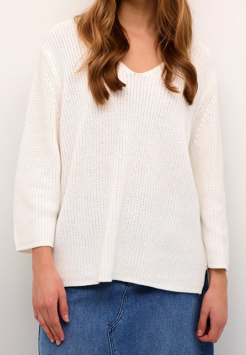 Femme portant un pull en maille blanc à manches trois-quarts et une jupe en denim bleu, debout devant un fond clair uni.