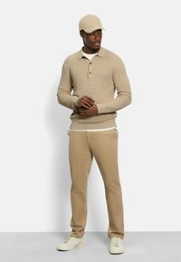 Beige Strick-Polohemd mit Kragen und drei Knöpfen, kombiniert mit hellbeigen Hosen und weißen Sneakers sowie einer passenden beige Kappe.