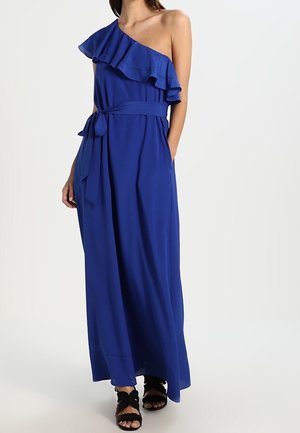 Vrouw draagt een koningsblauwe een-schouder jurk tot op de grond met volantdetails en een taillekoord, gecombineerd met zwarte sandalen met bandjes.
