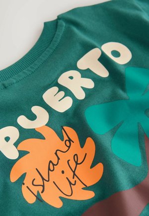 Grüner Baumwollpullover mit fettem weißem Text "PUERTO" und "island life" sowie orangefarbenen und türkisfarbenen Blattmotiven.