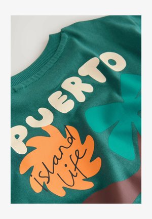 Zelena bombažna jopa z izrazitim belim napisom "PUERTO" in "island life" ter oranžnimi in turkiznimi listnatimi vzorci.