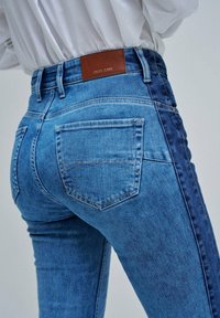 Primer plano de unos vaqueros de mezclilla azul con un panel lateral más oscuro, bolsillos traseros y una etiqueta de marca de cuero marrón, combinados con una camisa blanca metida por dentro.