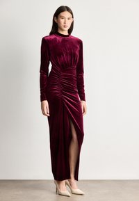 Μακρύ μανίκι φόρεμα σε μπορντώ βελούδο με πτυχώσεις και σκίσιμο ψηλά στο μηρό. Συνδυάζεται με μπεζ μυτερές γόβες. Λείο υφή.