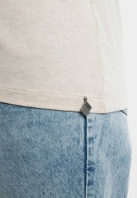 Lichtbeige kortefsleevem shirt van een gestructureerde stof, met een metalen label aan de zoom. Gecombineerd met lichtblauwe denim shorts met een vervaagd effect.