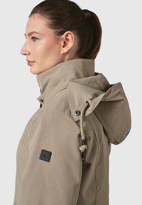 Bežs ūdensizturīgs žakets ar ribotu tekstūru, kapuci, regulējamiem aukliņām un melnu logo plāksteri uz pleca.