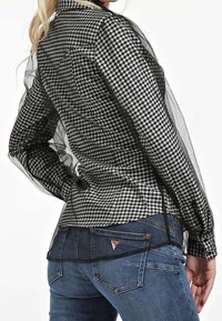 Blouse à carreaux noir et blanc avec des manches transparentes et un ourlet noir uni. Associée à un jean bleu avec un accent logo sur la poche.