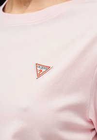 Ανοιχτό ροζ μπλουζάκι με ένα μικρό τριγωνικό λογότυπο Guess στο αριστερό στήθος, με κόκκινες, άσπρες και μπλε αποχρώσεις.